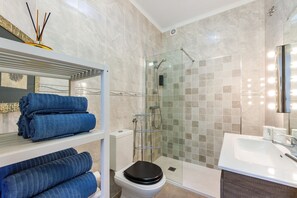 Une salle de bain avec une douche à parois de verre, un lavabo blanc et des étagères où sont pliées des serviettes.