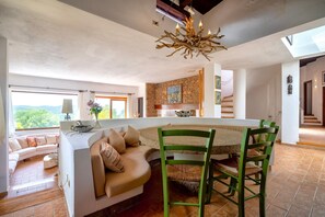 A modern living room with a curved sofa, a dining area with a green chair, a chandelier, and a stone wall. - Villa 'Querida' with Sea View, Private Pool and Private Terrace (Costa Paradiso)