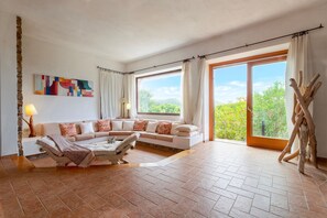A spacious living room with a large window, a sofa, a coffee table, and a lamp. - Villa 'Querida' with Sea View, Private Pool and Private Terrace (Costa Paradiso)