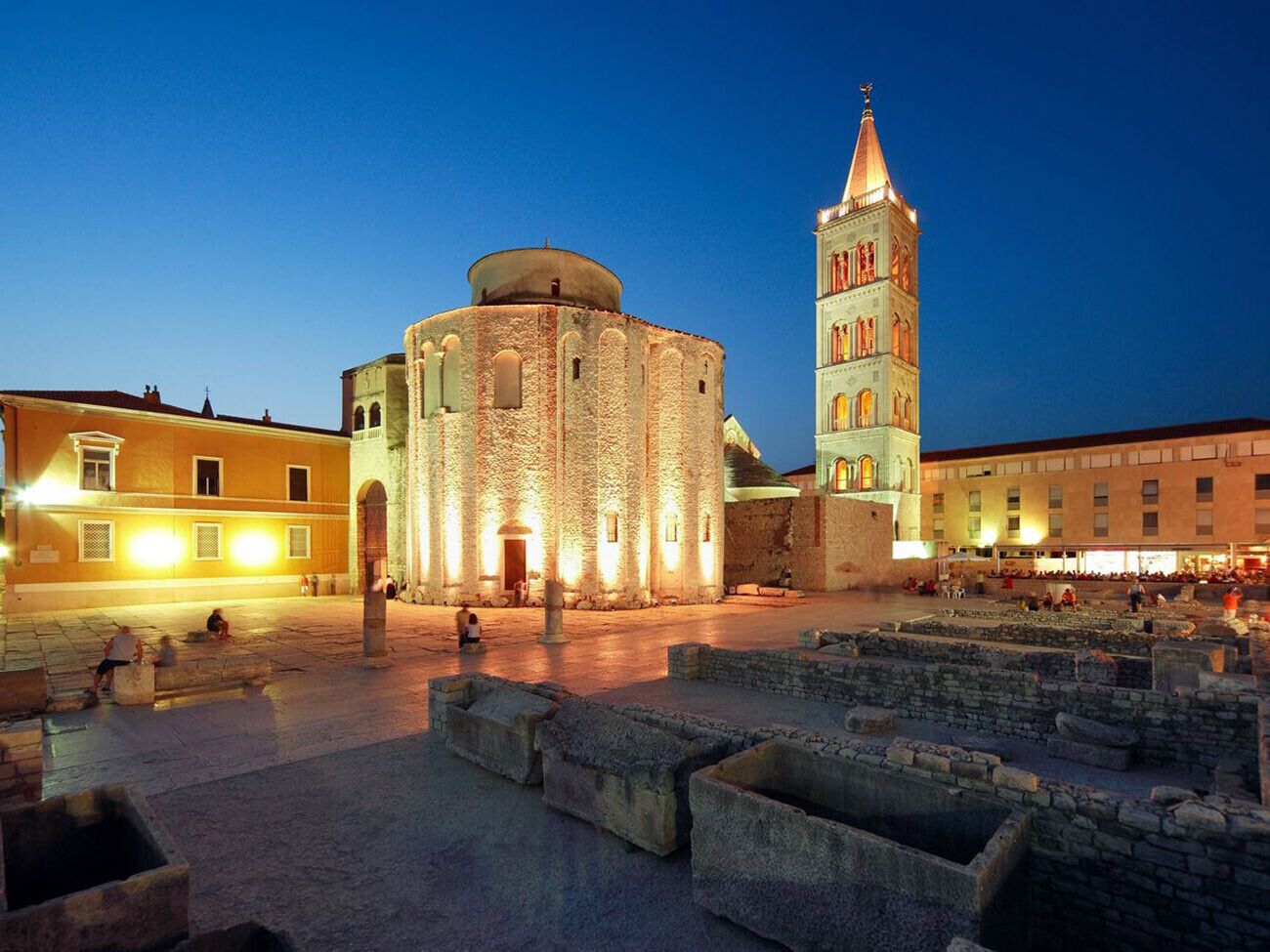 A historic square with a round building and a tall tower, surrounded by stone ruins and modern buildings.