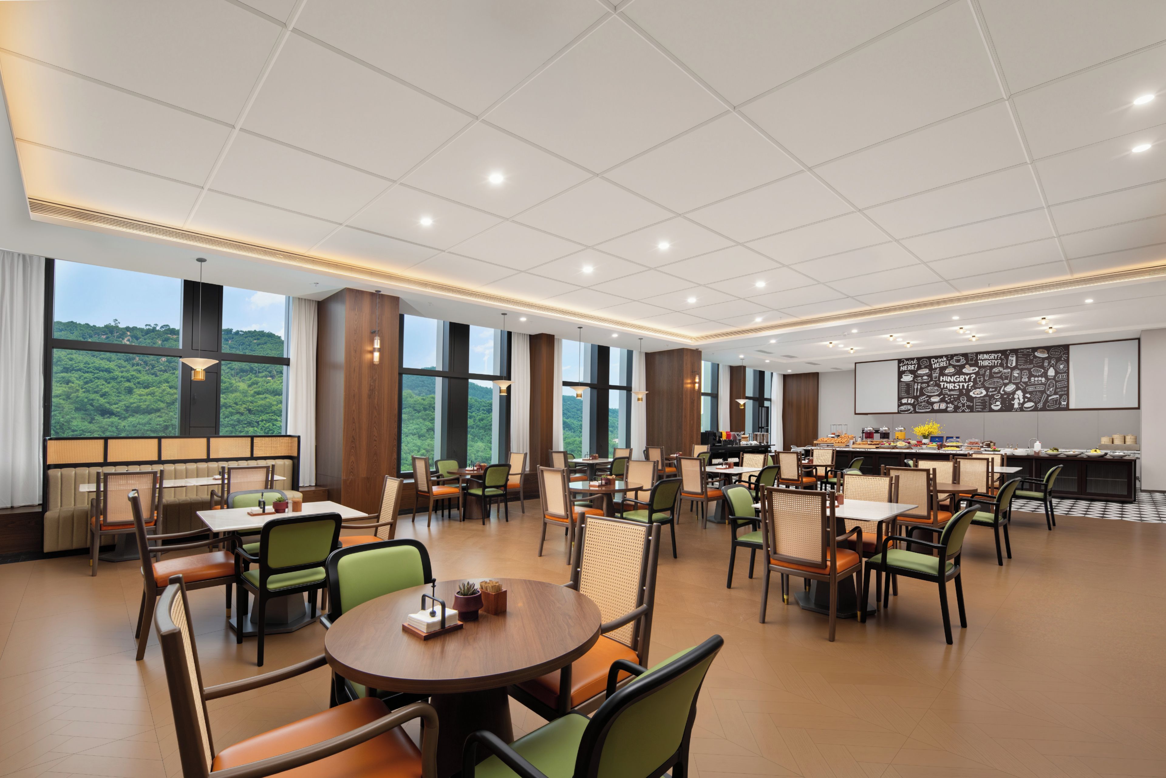Un vaste espace dédié à la salle à manger, avec de nombreuses tables et chaises, de grandes fenêtres offrant une vue sur la verdure, et un coin cuisine doté d’un tableau noir servant de menu.