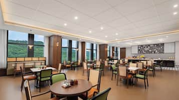 Un vaste espace dédié à la salle à manger, avec de nombreuses tables et chaises, de grandes fenêtres offrant une vue sur la verdure, et un coin cuisine doté d’un tableau noir servant de menu.