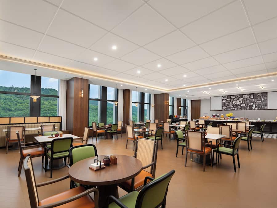 A spacious dining area with multiple tables and chairs, large windows offering a view of greenery, and a kitchen area with a chalkboard menu.