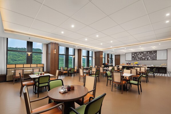 A spacious dining area with multiple tables and chairs, large windows offering a view of greenery, and a kitchen area with a chalkboard menu.