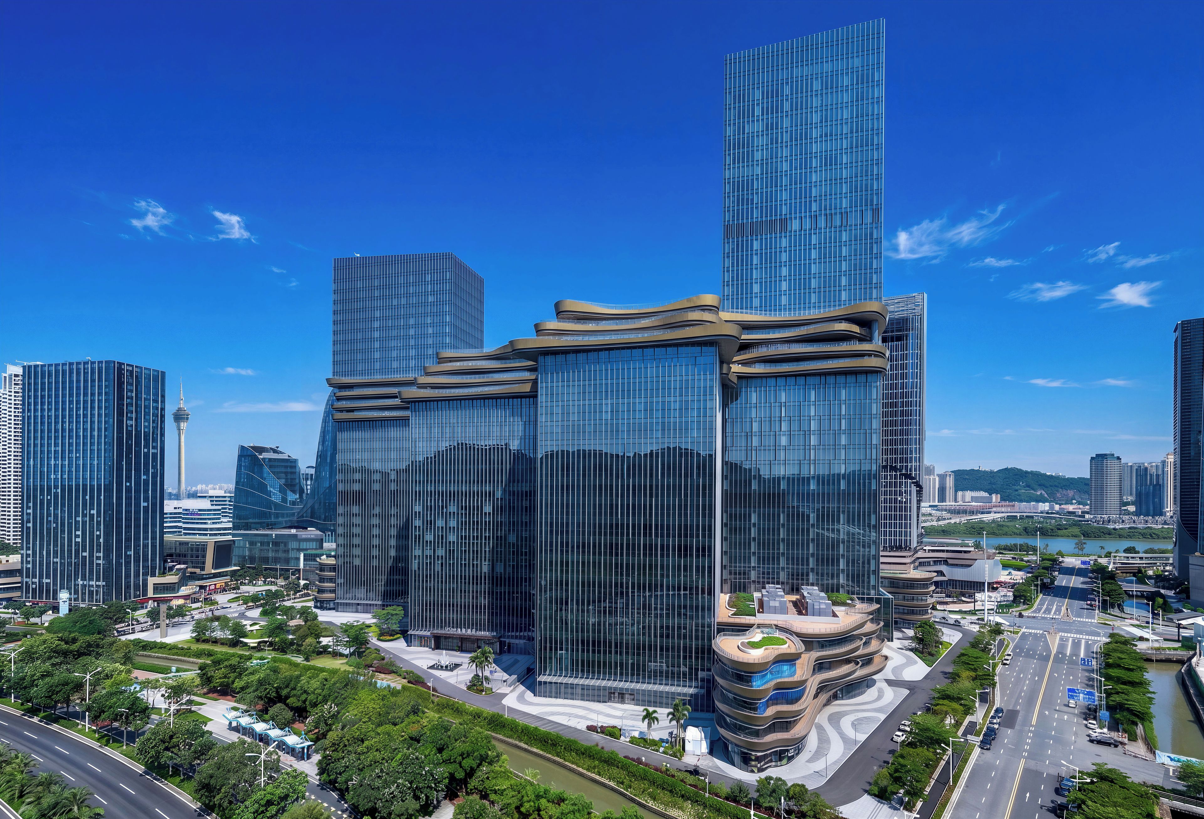 A modern high-rise building with a curved glass facade, surrounded by a well-maintained urban park and a busy road.