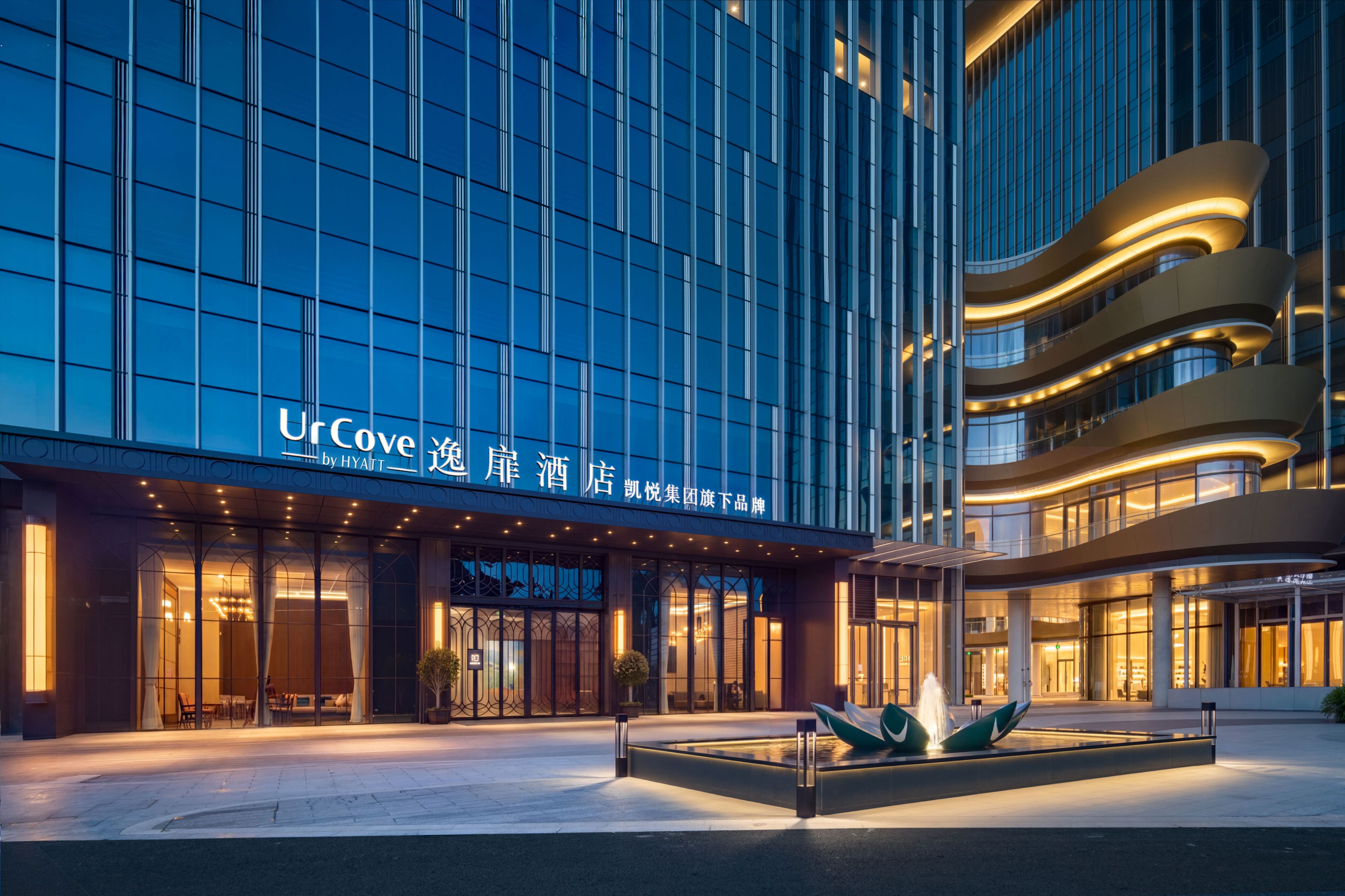 A modern hotel building with a glass facade, a well-lit entrance, and a landscaped area with a water feature.