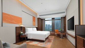 Habitación de hotel con cama, escritorio, silla, televisión y un amplio ventanal con vista a la ciudad.