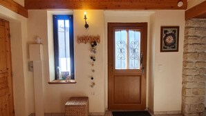 Una stanza con struttura in legno, una porta, una piccola panca, una lampada a parete, una finestra con tende e un muro in pietra.