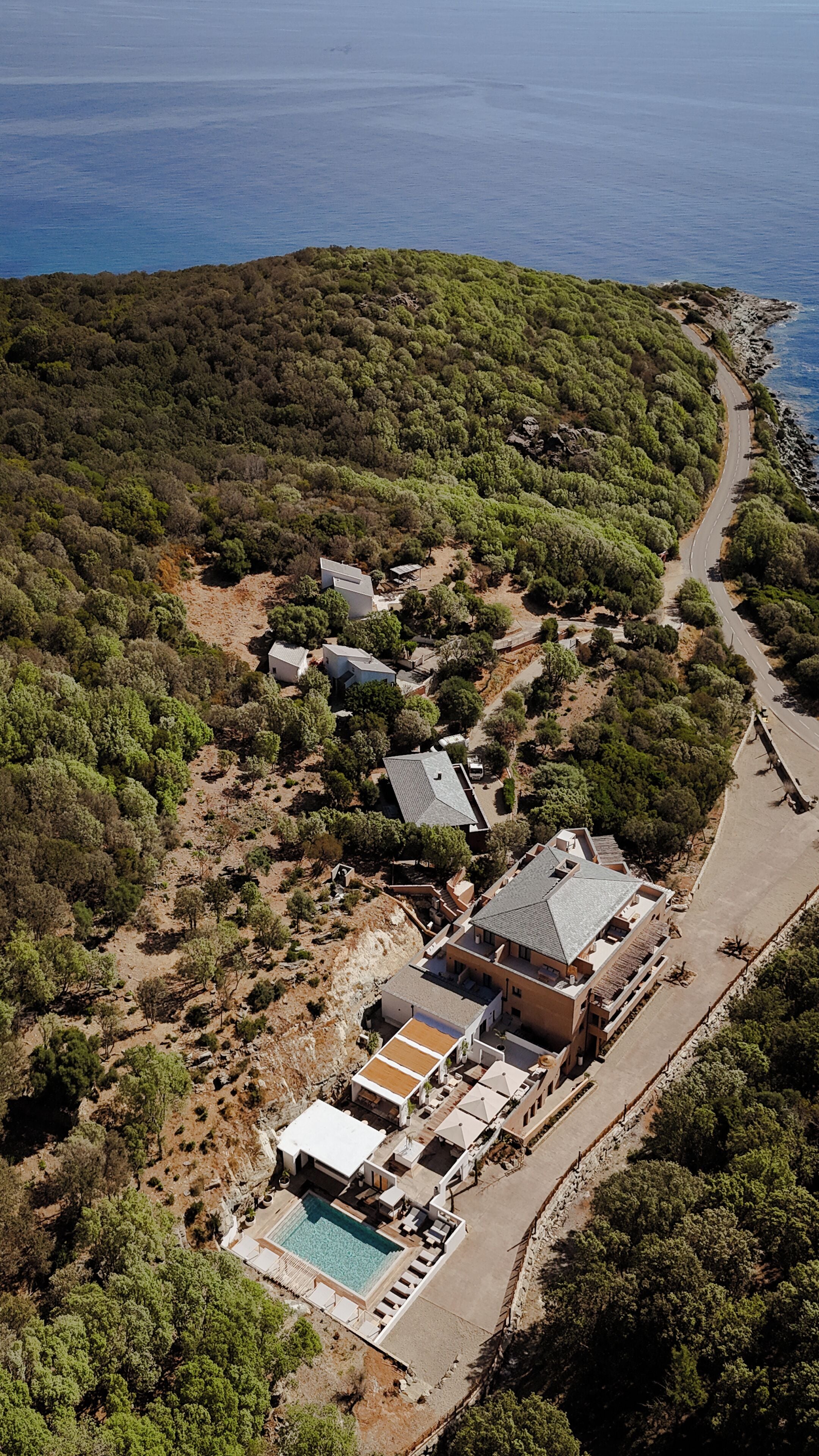 An aerial view of a coastal property with a swimming pool, multiple buildings, and a road leading to the sea.
