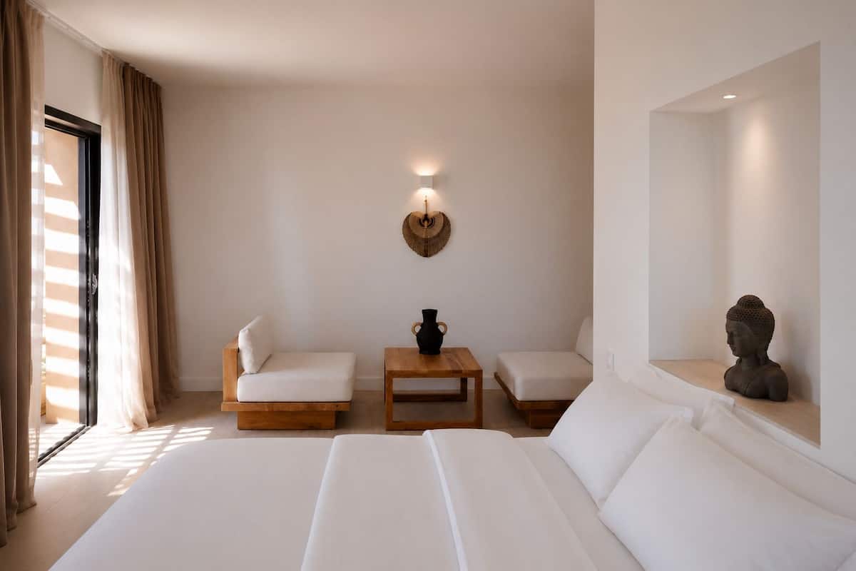 A hotel room with a large bed, two white sofas, a small wooden table, a wall-mounted lamp, and a bust sculpture.