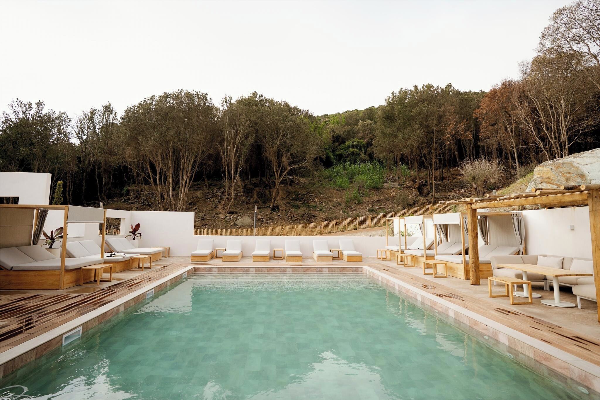 A pool area with lounge chairs and wooden tables, surrounded by trees.