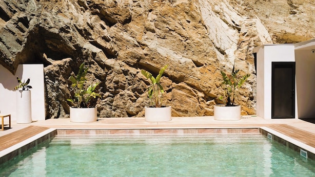 A swimming pool with clear water, surrounded by white planters with plants, against a rocky cliff backdrop. - Hotel Casamore (Meria)