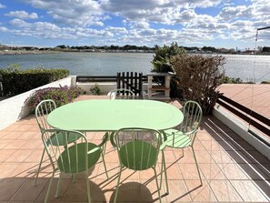 A patio with a round table and chairs, a black fence, and a view of a body of water and buildings in the distance. - Cottage with air conditioning, lake view - 5 guests (Le Barcarès)