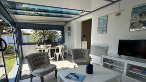 A modern living room with a glass-enclosed patio, white furniture, a flat-screen TV, and a dining area visible through the glass. - House near the sea - 5 guests (Noirmoutier-en-l'Île)
