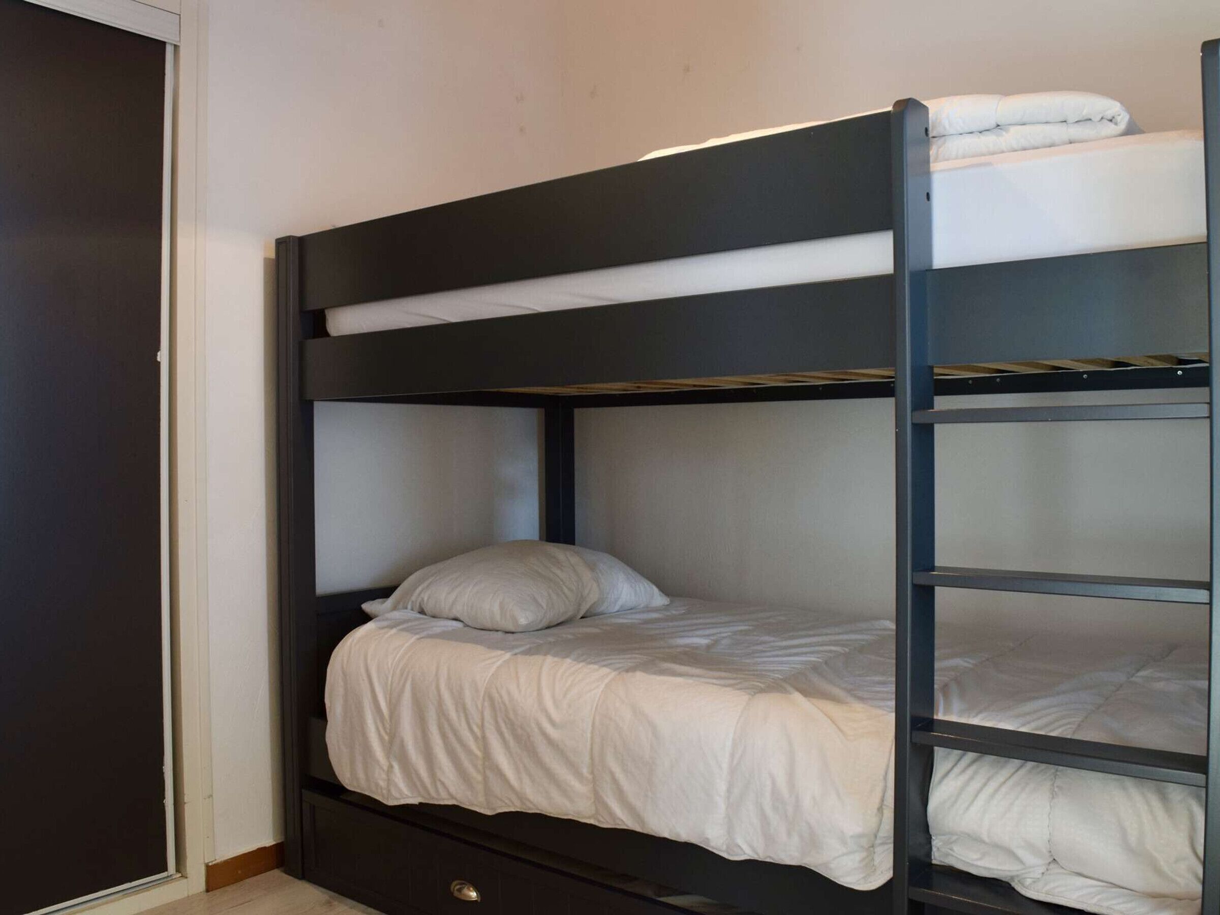 Un lit superposé avec un matelas et un oreiller blancs, une tête de lit en bois et une armoire de couleur foncée, située à gauche.