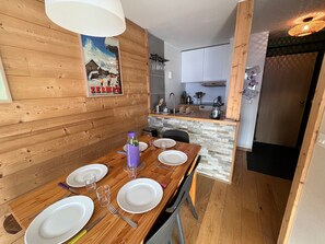 A wooden dining table with plates, cutlery, and glasses, a kitchen area with cabinets, and a mounted picture of a snowy scene. - Avoriaz - Apartment for 5 ppl, village and ski area view (Avoriaz)
