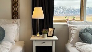Un dormitorio con dos camas, una mesita de noche con lámpara, un cuadro enmarcado y una ventana con vistas a montañas nevadas.