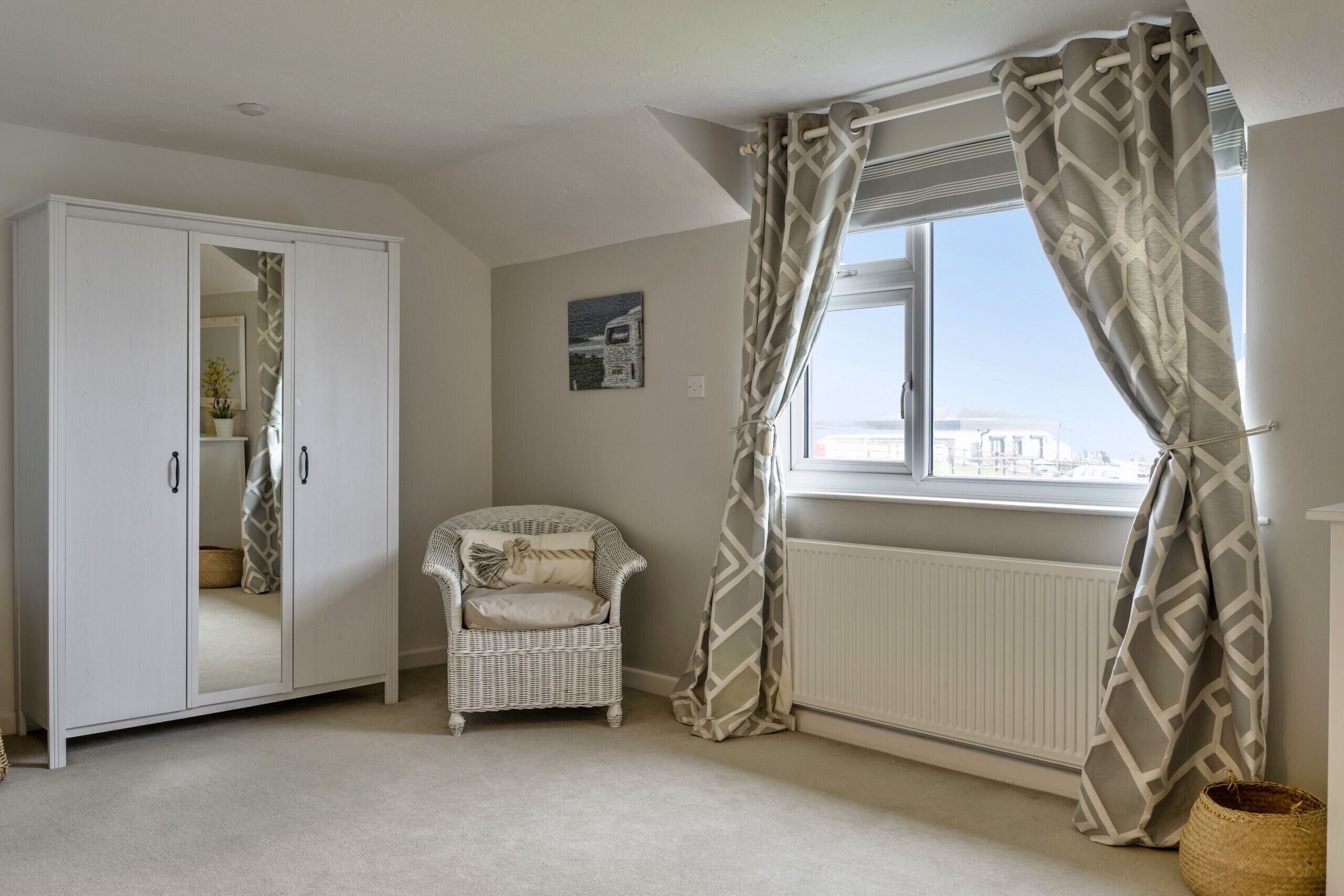 A room with a wardrobe, a wicker chair, a radiator, and a window with patterned curtains.