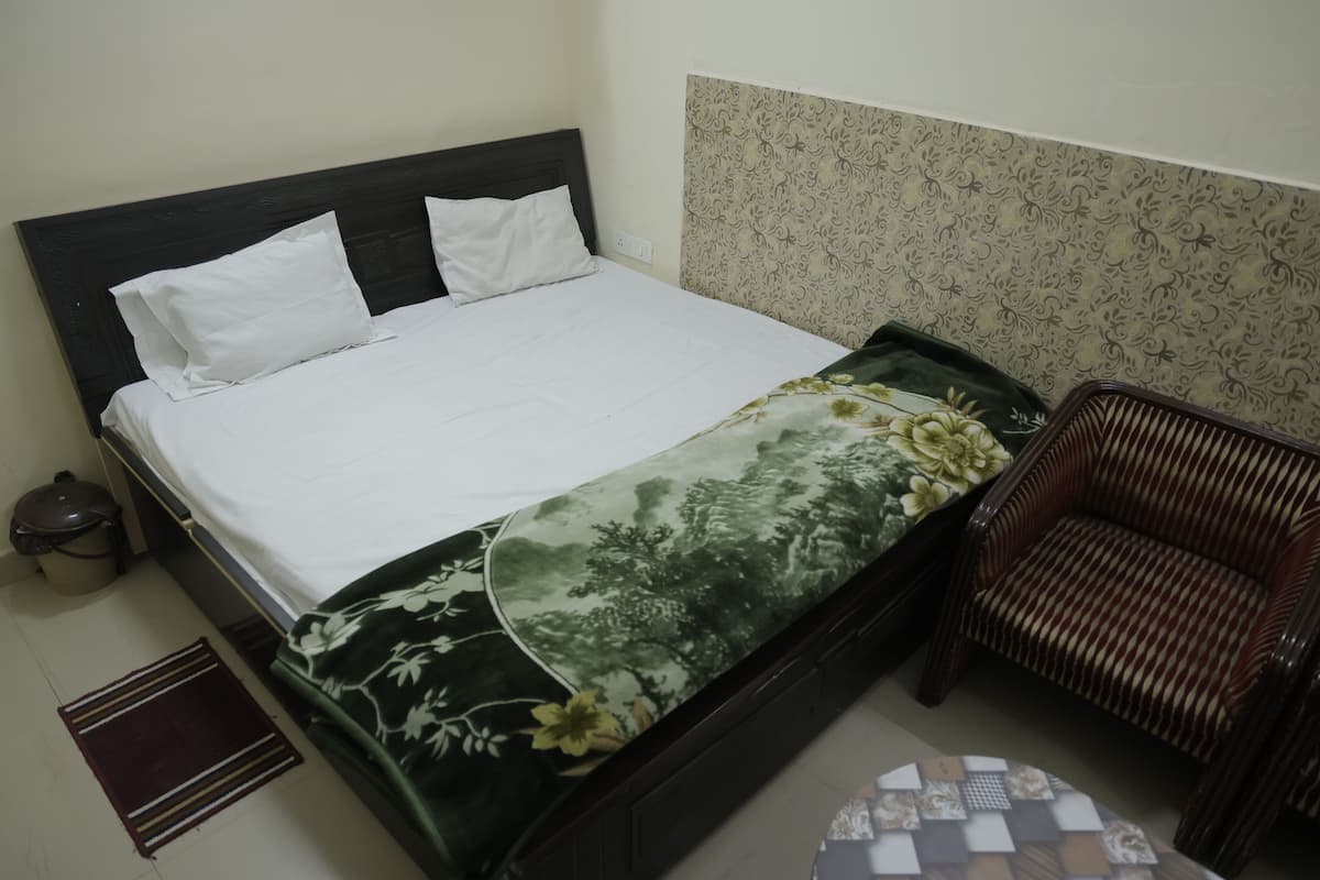 A bed with white linens, a floral-patterned bedspread, a dark wooden headboard, a patterned wall, a wicker chair, and a small round table with a checkered tablecloth.