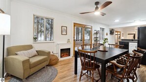 Un salon chaleureux avec une cheminée, un coin repas avec des chaises en bois et une cuisine dotée d’un réfrigérateur noir.