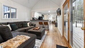 Un salon moderne avec un canapé, un pouf et une table basse. Il y a également un coin cuisine avec une table à manger et des chaises. La pièce est dotée d’une grande porte-fenêtre coulissante qui donne sur une terrasse extérieure.