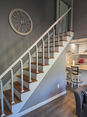 A staircase with wooden steps and white railings, a kitchen area with bar stools, and a wall clock. - Grand opening to gorgeous Asheville house! (Asheville)