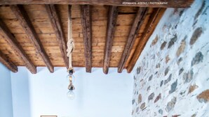 A room with a wooden ceiling, stone wall, bed with patterned bedding, a bench, a small table with decorative items, and framed pictures on the wall. - Apartment 'The Balcony Of Bougainvillea' with Sea View, Wi-Fi and Air Conditioning (Ano Syros)