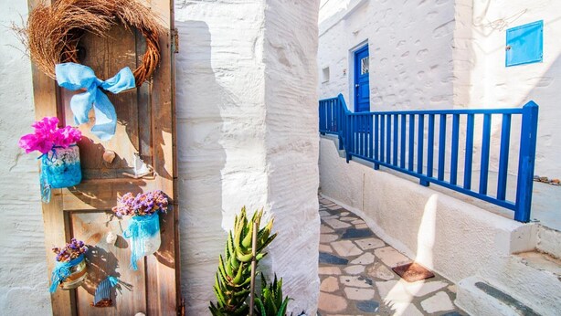 A white-walled building with a wooden door adorned with a wreath and flowers, a blue railing, and a stone-paved path. - Apartment 'The Balcony Of Bougainvillea' with Sea View, Wi-Fi and Air Conditioning (Ano Syros)