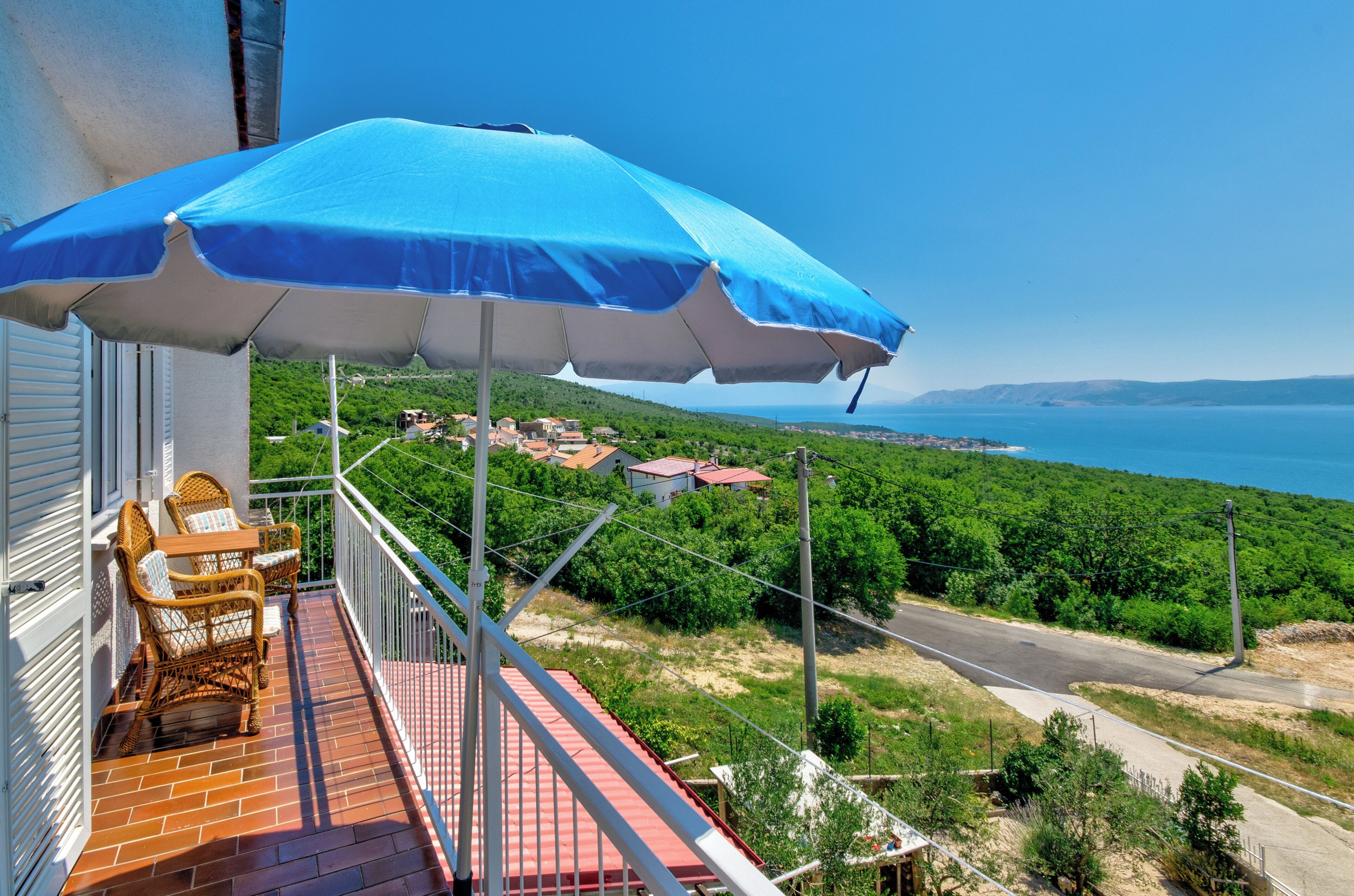 Een balkon met rieten meubilair, een blauwe parasol en uitzicht op een kustlandschap.