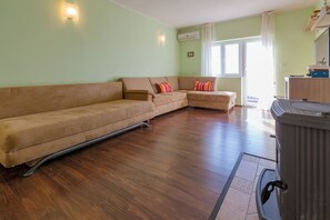Ein Wohnzimmer mit einem beigefarbenen Sofa, Holzboden, Heizkörper und einem Fenster mit Vorhängen.