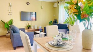 Moderni olohuone, jossa on litteänäyttöinen televisio, sohvia, ruokapöytä, jonka päällä on lautasia ja laseja, sekä maljakko kukkineen.