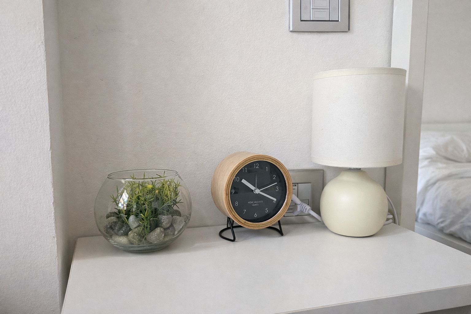 A bedside table with a clear glass fishbowl containing plants, a wooden analog clock, and a white table lamp.