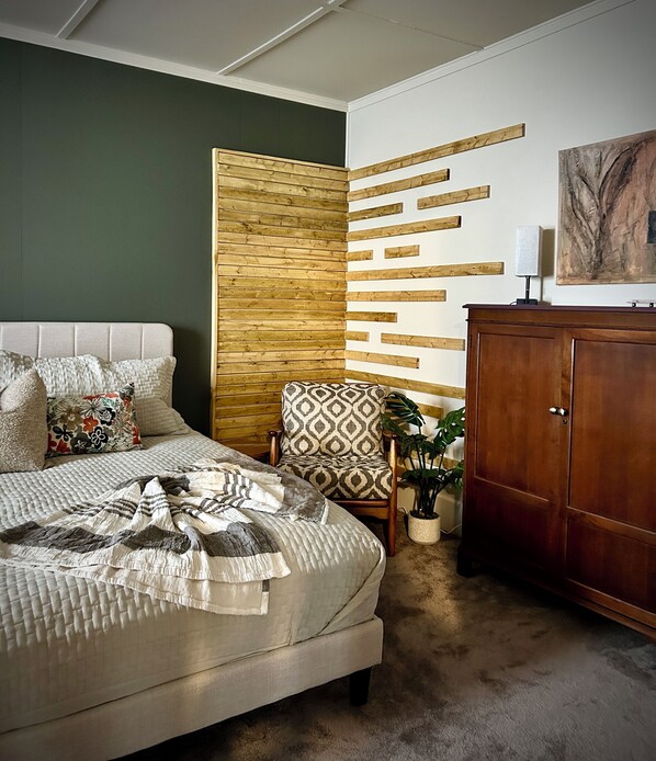 A bedroom with a bed, a patterned armchair, a wooden cabinet, and a wall-mounted light fixture.