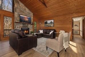 A log cabin interior with a stone fireplace, leather sofas, a television, and a large window overlooking trees. - A Change in Altitude | GAME ROOM | HOT TUB | Hiking| Library| Sleeps 12 (Deep Gap)