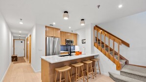 A modern kitchen with a central island, bar stools, wooden cabinets, stainless steel appliances, and a staircase with wooden railings. - New Listing! Walkable Frisco Condo | Near Breck & Copper | Pool + Hot Tub (Frisco)