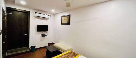 A hotel room with a bed, a desk, a chair, a television, an air conditioning unit, and a ceiling fan.