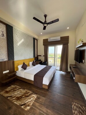 A hotel room with a bed, a ceiling fan, an air conditioning unit, a TV, and a balcony with a view. - Kapish Farms by Kavya International (Udaipur)