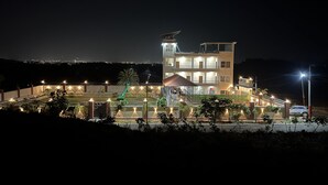 A multi-story hotel with a prominent rooftop structure, a well-lit entrance, and a landscaped garden area. - Kapish Farms by Kavya International (Udaipur)