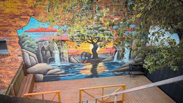 Een muurschildering van een waterval en boom op een bakstenen muur, een houten terras en een metalen reling.