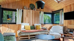 Un intérieur de chalet chaleureux avec un canapé, un fauteuil et une table ronde, de grandes fenêtres avec des rideaux et un téléviseur fixé au mur.