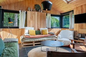 Interior acogedor de una cabaña con sofá, sillón y una mesa redonda, ventanas amplias con cortinas y un televisor montado.
