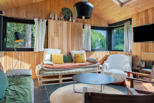 Un intérieur de chalet chaleureux avec un canapé, un fauteuil et une table ronde, de grandes fenêtres avec des rideaux et un téléviseur fixé au mur.