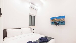 Hotelkamer met een bed, een raam, een airconditioner en een aan de muur bevestigde televisie waarop een stadsbeeld te zien is.