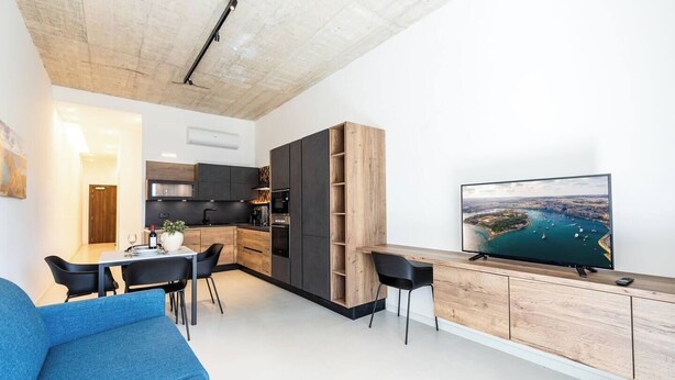 Een moderne keuken met donkere kasten, een zithoek met een blauwe bank en een televisie waarop een uitzicht op de kust te zien is.