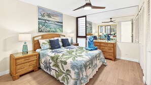 Quarto com cama, mesas de cabeceira, ventoinha no teto, um quadro grande, espelho e janela.