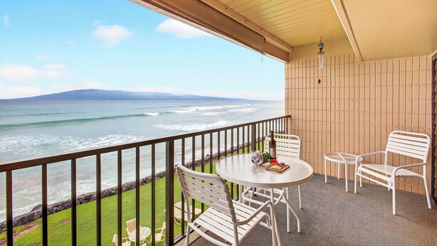 Uma varanda com vista para o oceano, uma mesa redonda com cadeiras e uma mesinha com uma garrafa e copos.