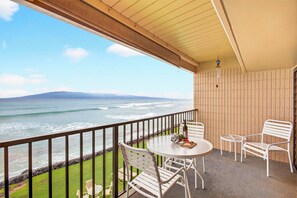 A balcony with a view of the ocean, a round table with chairs, and a small table with a bottle and glasses. - Simple Condo Great View (Lahaina)