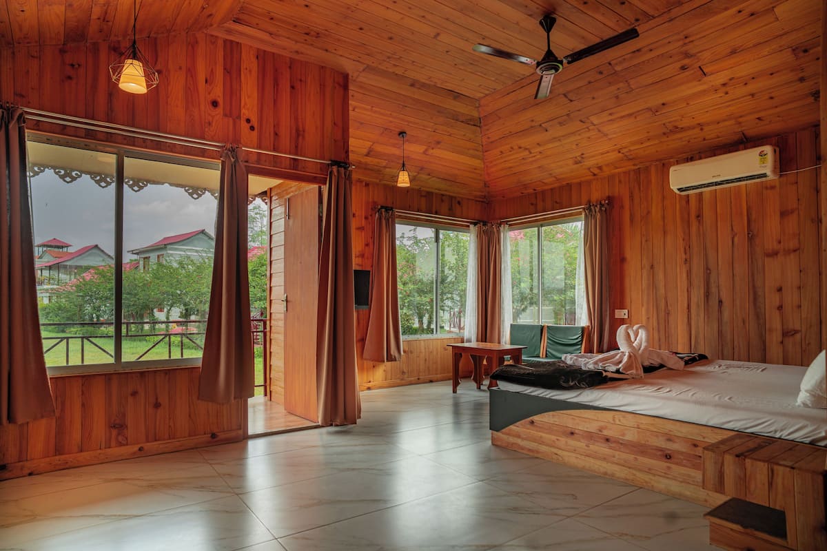 A wooden cabin room with a bed, a table, and a view of a garden through large windows.