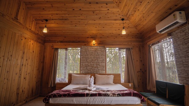 A wooden cabin bedroom with a bed, two hanging lights, a bench, and an air conditioning unit. - Resort Bihanga (Mal)