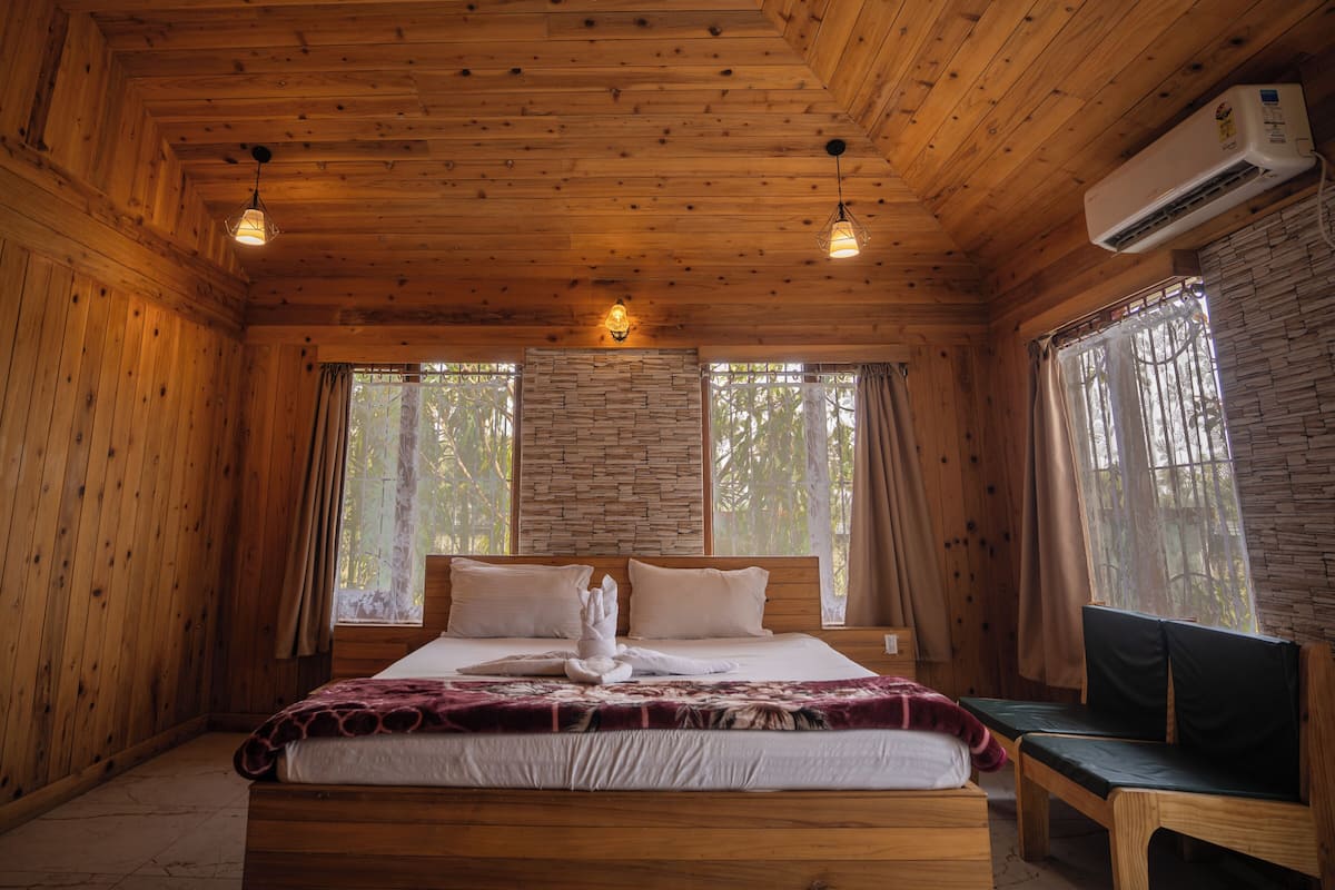 A wooden cabin bedroom with a bed, two hanging lights, a bench, and an air conditioning unit.